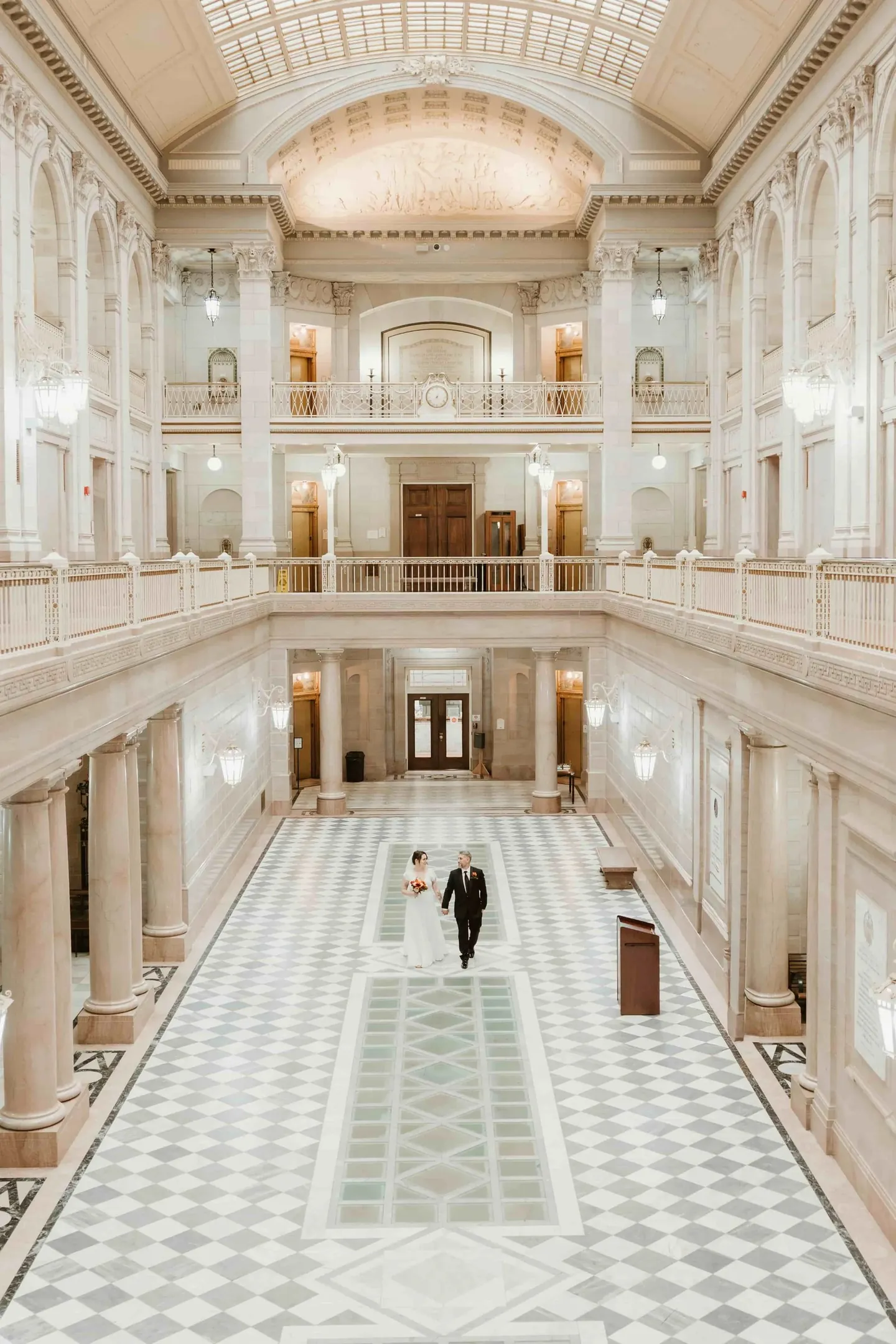 Amber and Marco stand small at the center of the marble atrium beneath stacked balconies