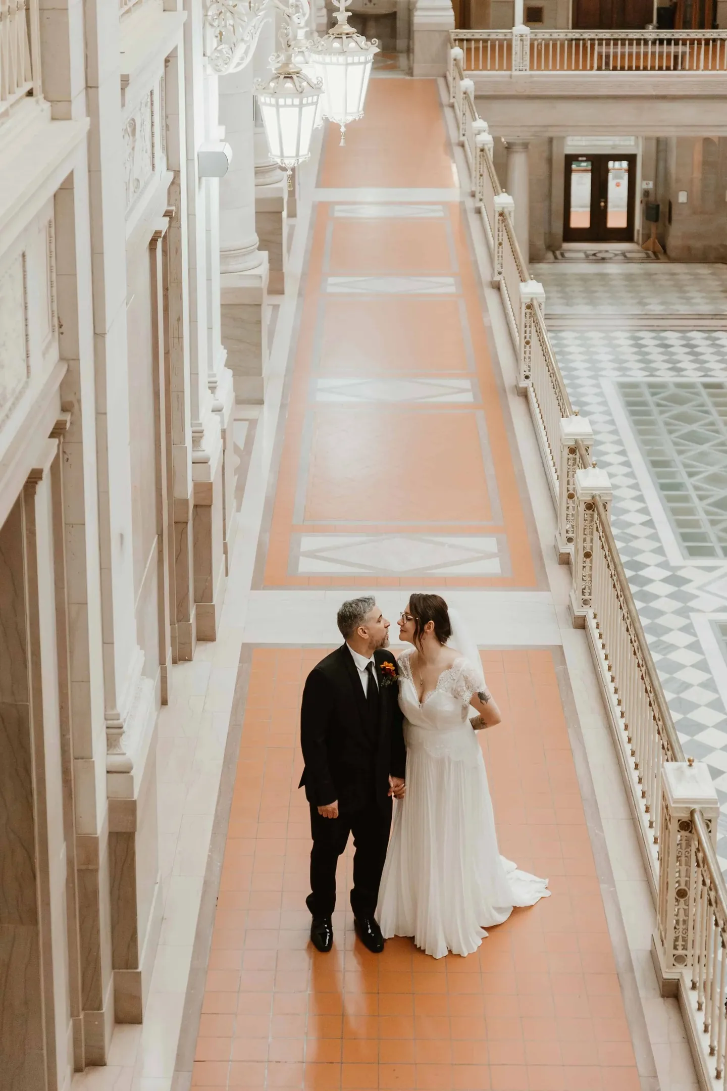 Amber and Marco stand in a long terracotta corridor lined with lamps, seen from above