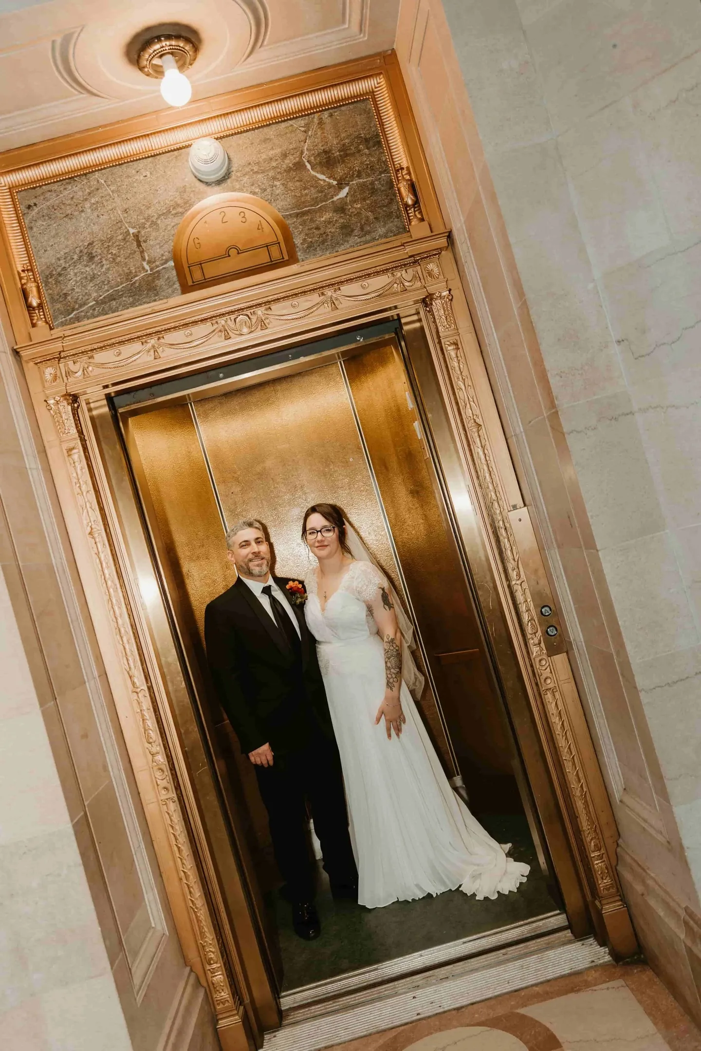 Amber and Marco stand inside an ornate brass elevator with its doors open