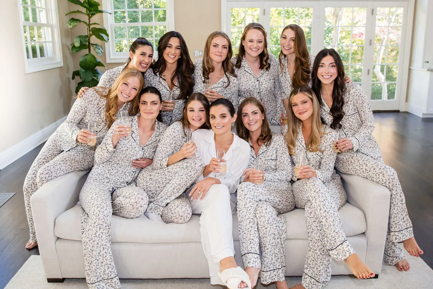 Bridesmaids in matching printed pajamas crowd onto a white sofa, laughing in soft window light