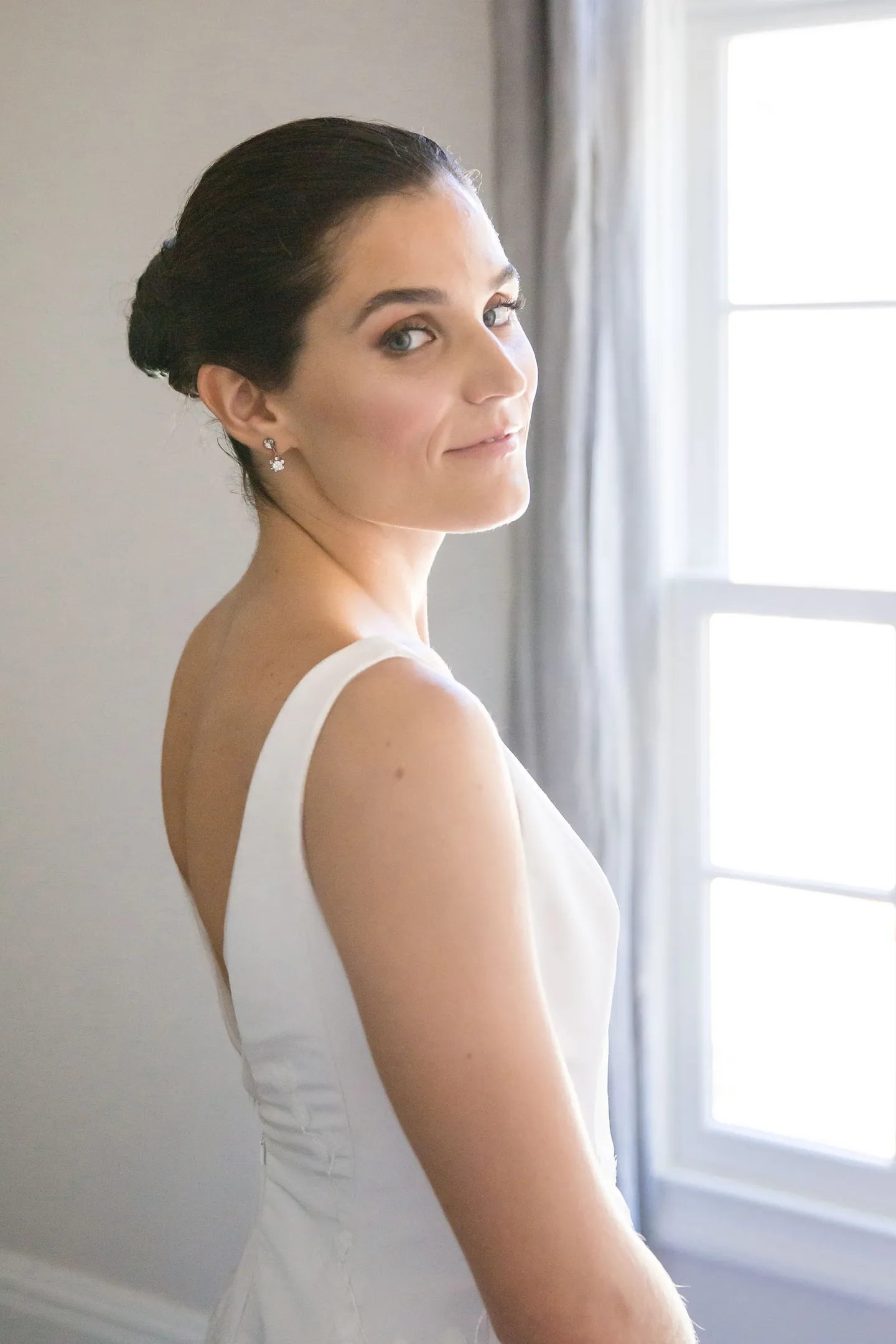 The bride in a sleeveless gown glances over her shoulder beside a tall white window