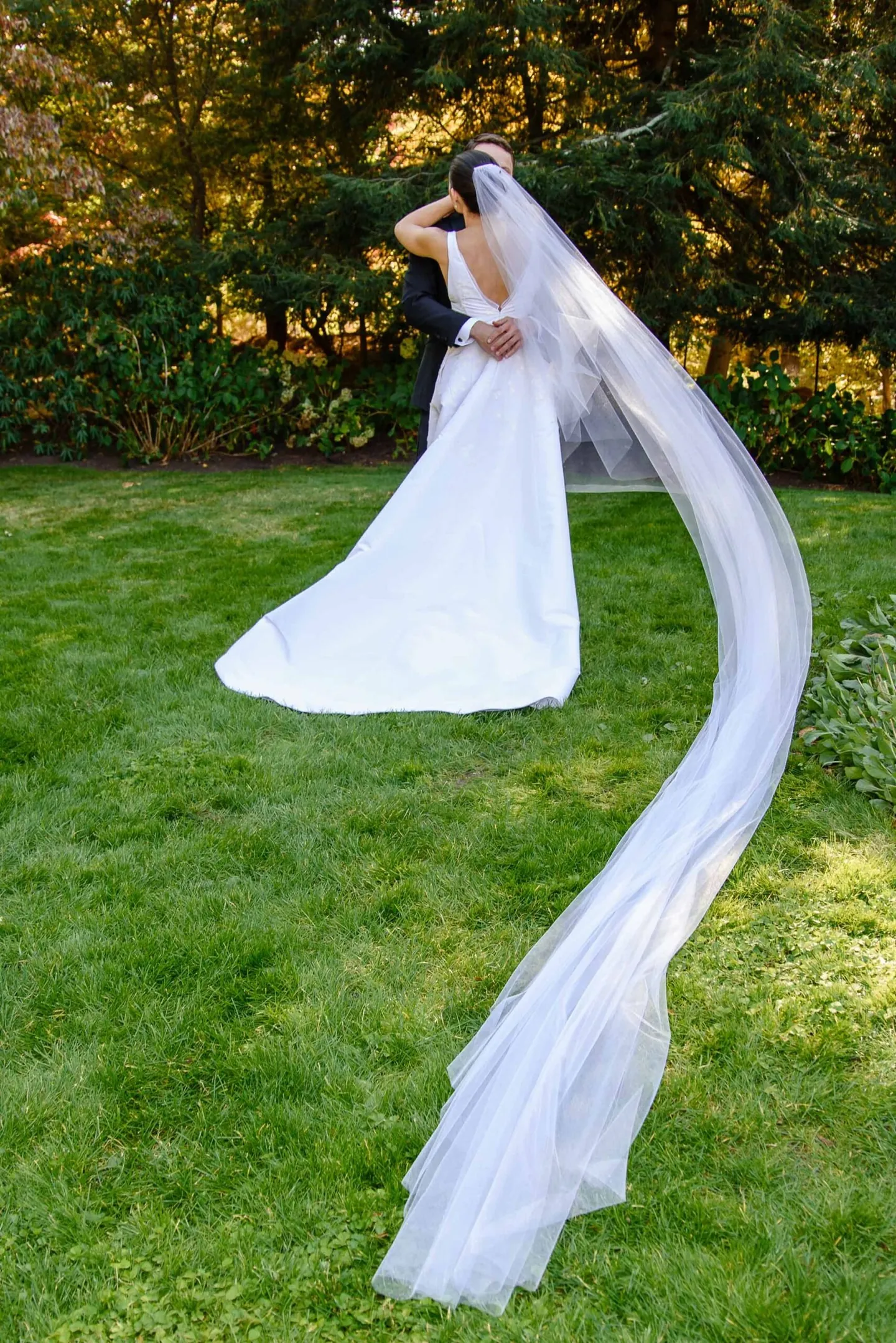 The bride's long veil streams sideways across the green lawn in a strong breeze