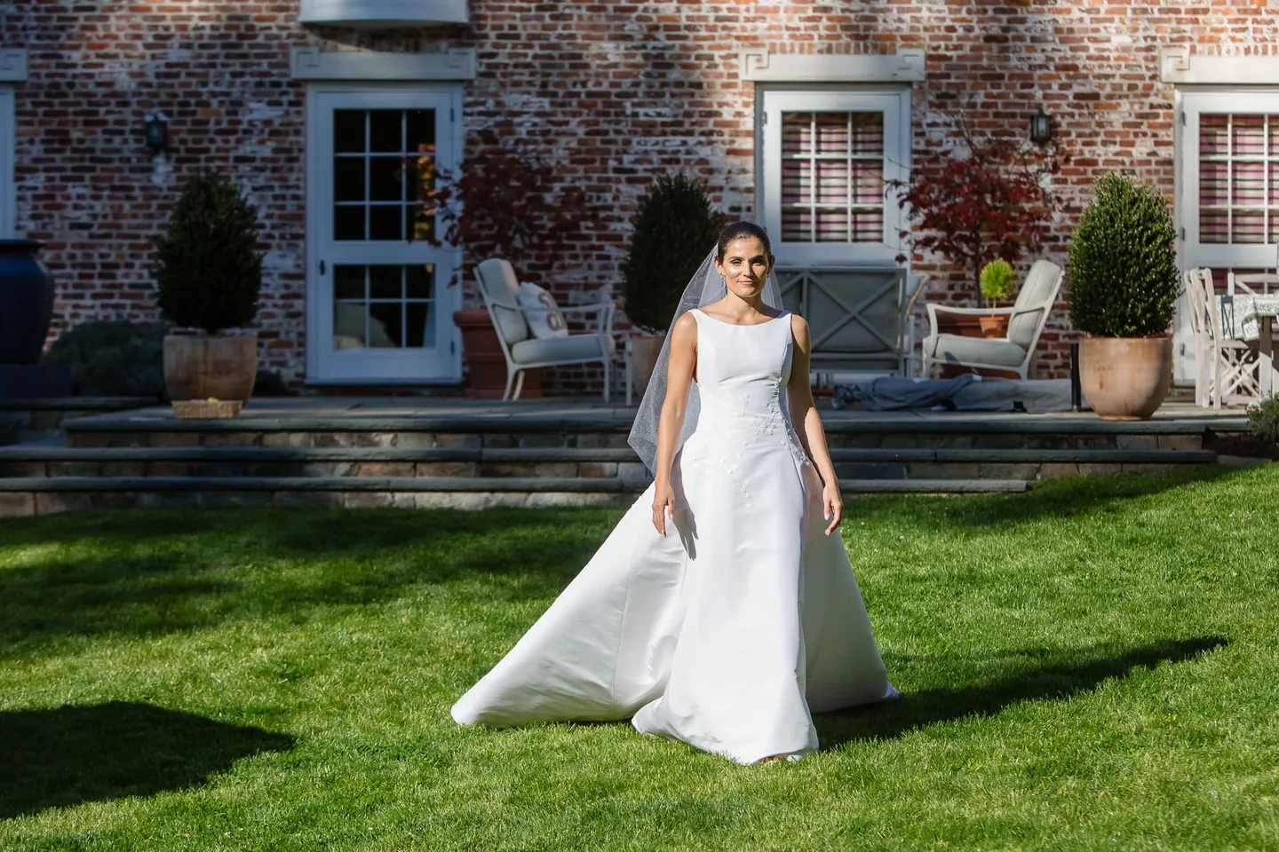 The bride walks across the lawn in front of a stone house with dark shutters