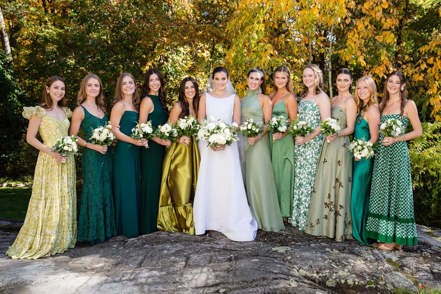 Bridesmaids in mismatched green gowns stand on a rock ledge with the bride in the middle