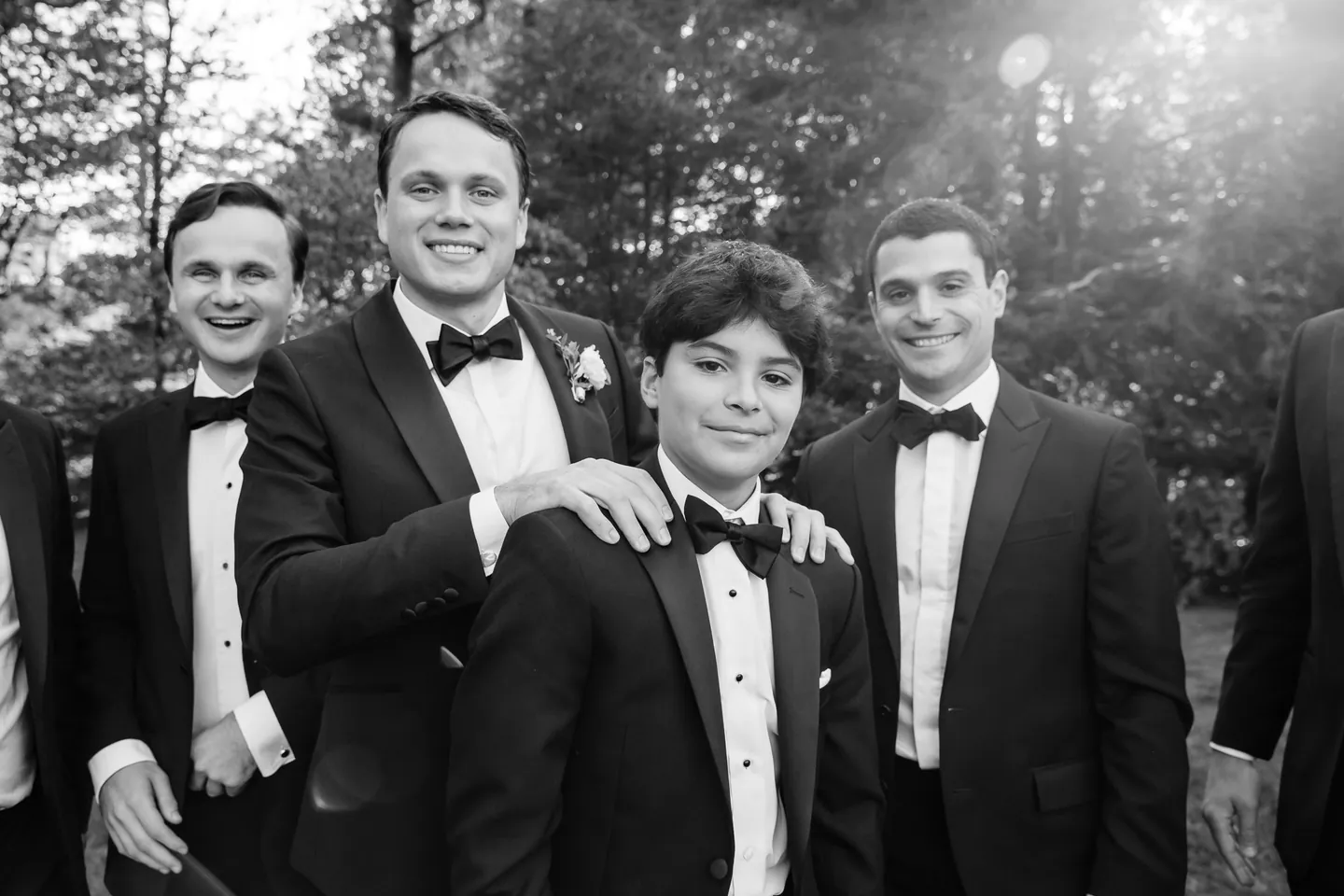 Groomsmen in tuxedos grin with hands on shoulders, a young guest in front, in black and white