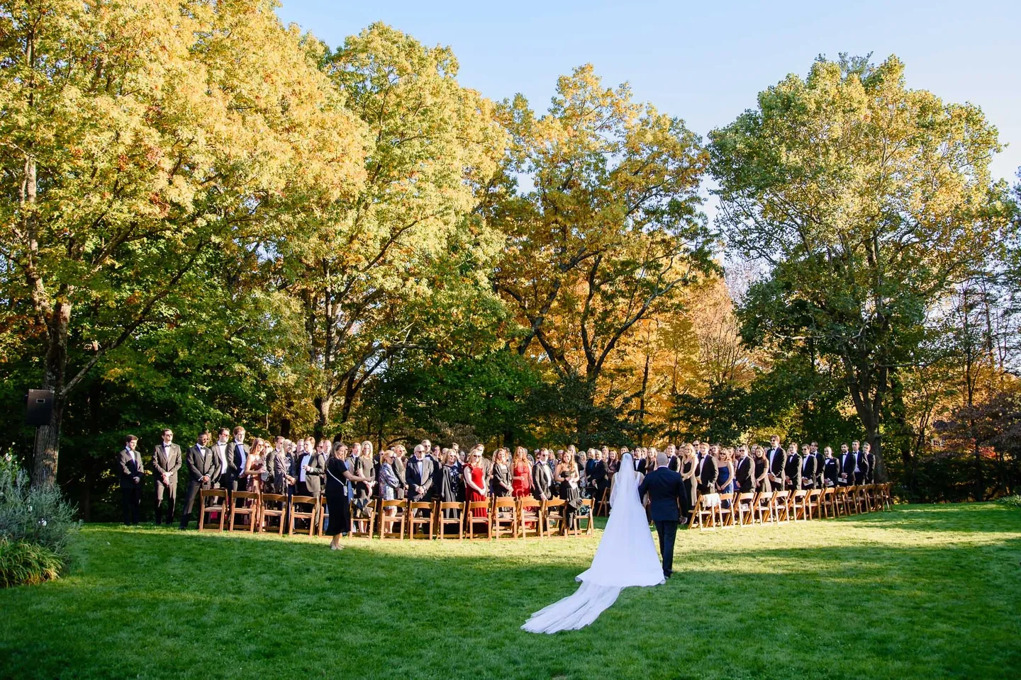 The bride and her father walk toward rows of standing guests under yellowing trees