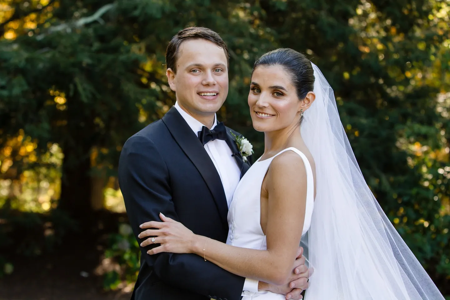 Angie and Matt hold each other and smile at the camera against dark evergreens