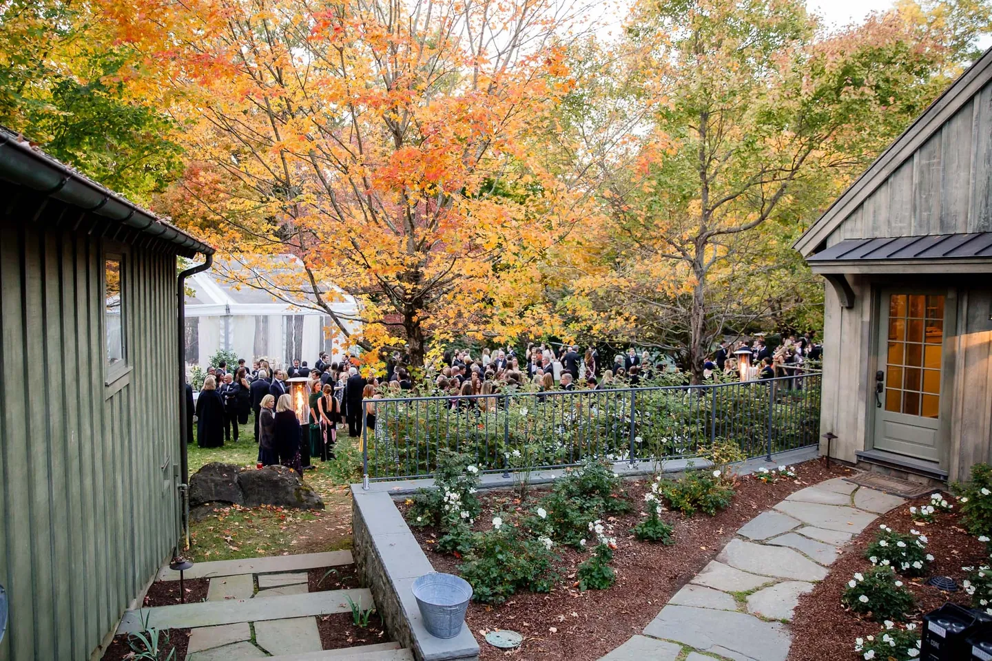 Guests gather along a stone path between gray barns as the trees turn orange