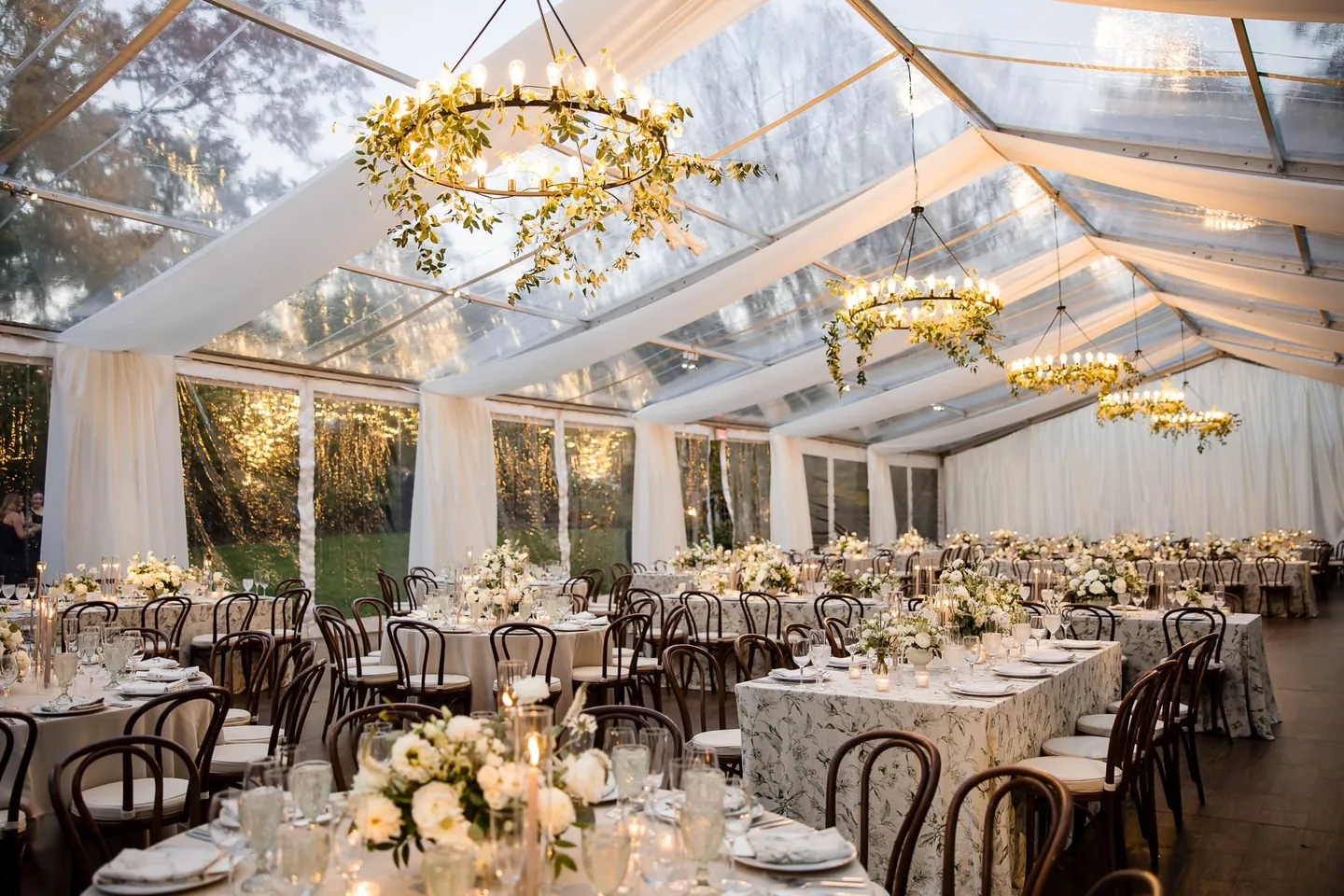 Chandeliers wrapped in greenery hang inside a clear-top tent set for dinner