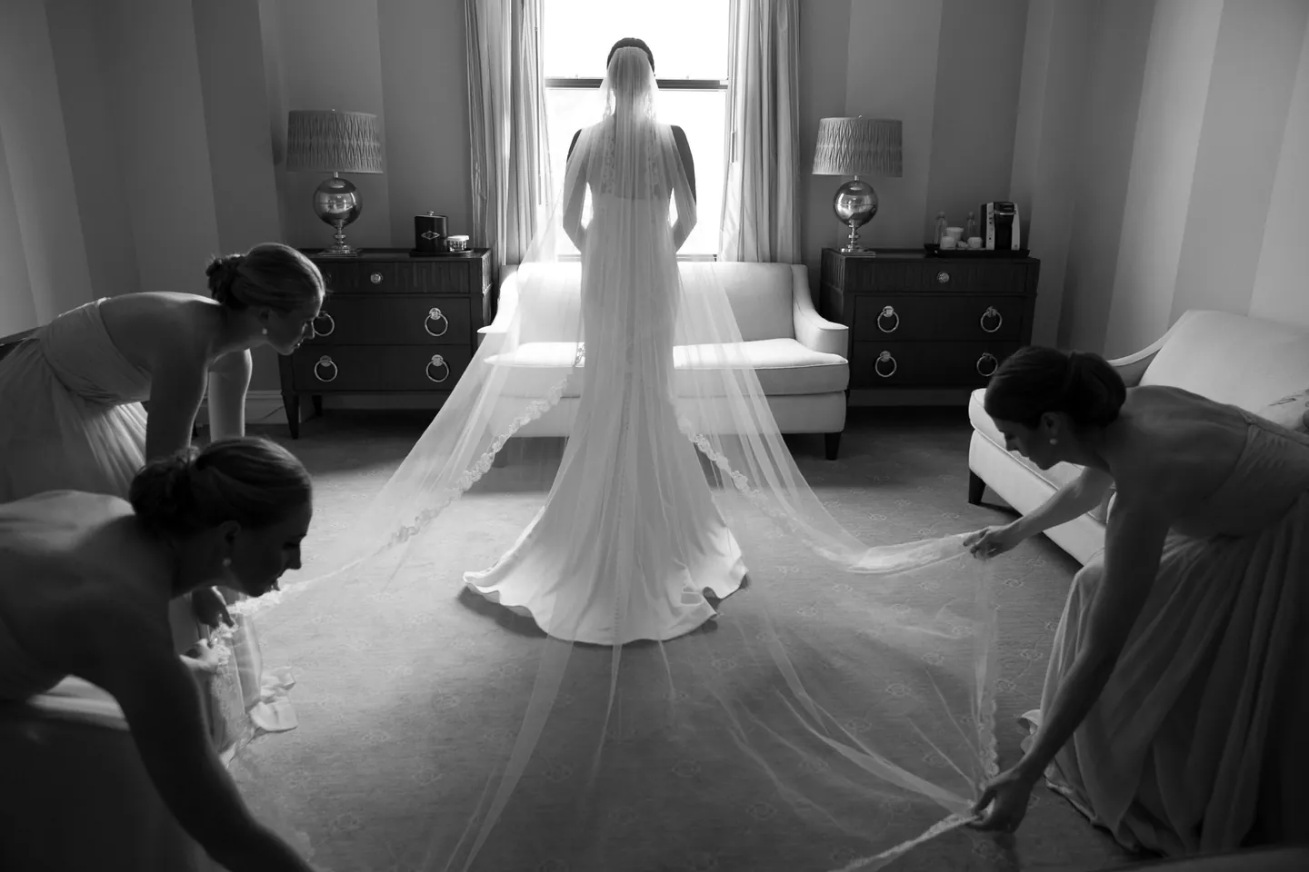 Bridesmaids kneel to arrange the bride's long train on the carpet, in black and white
