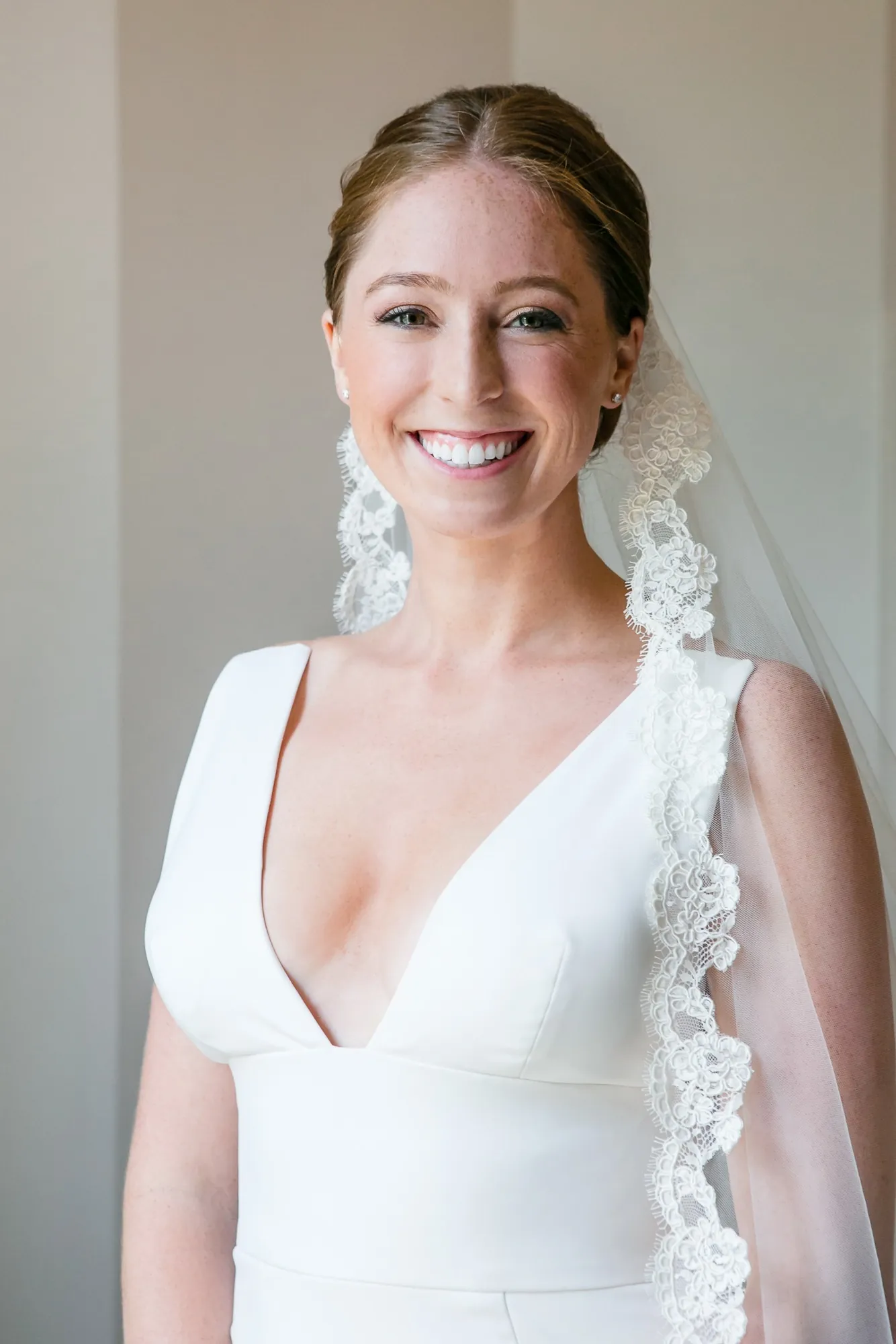 The bride in a deep V gown and lace-edged veil smiles beside a window