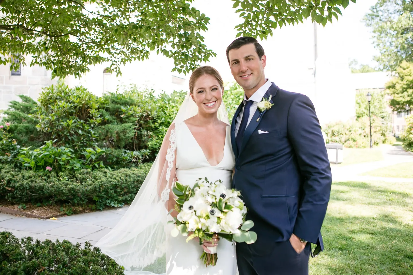 Anna and John smile together under a leafy tree, her bouquet full of white anemones