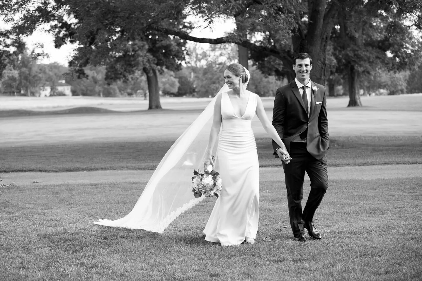 Anna and John walk across the open lawn under big trees, in black and white