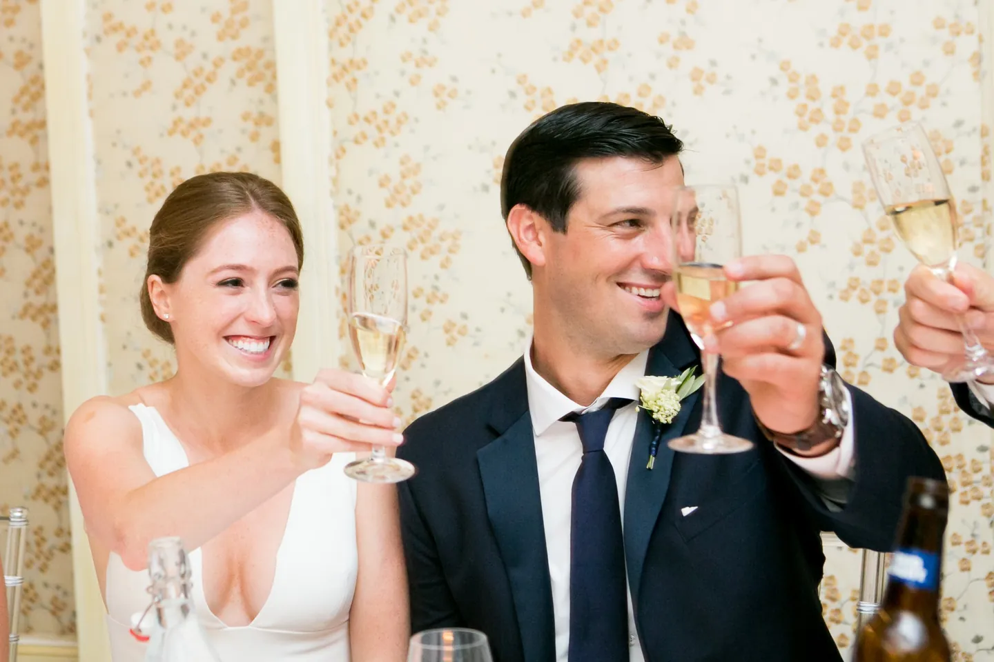 Anna and John raise champagne glasses and laugh at the head table