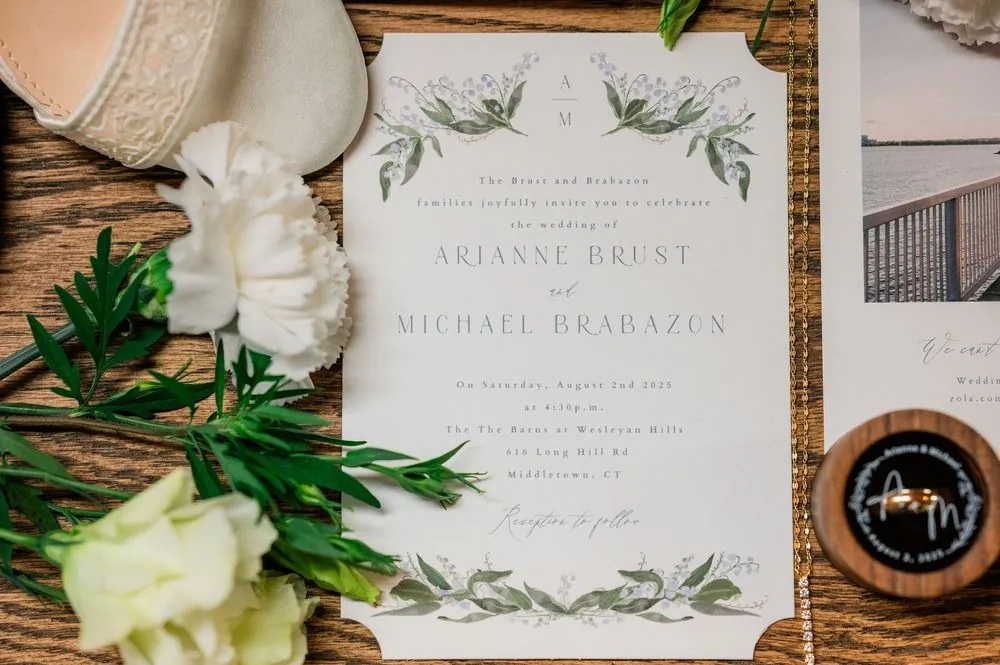 Invitation suite with greenery watercolor on a wood table beside white roses and wedding bands