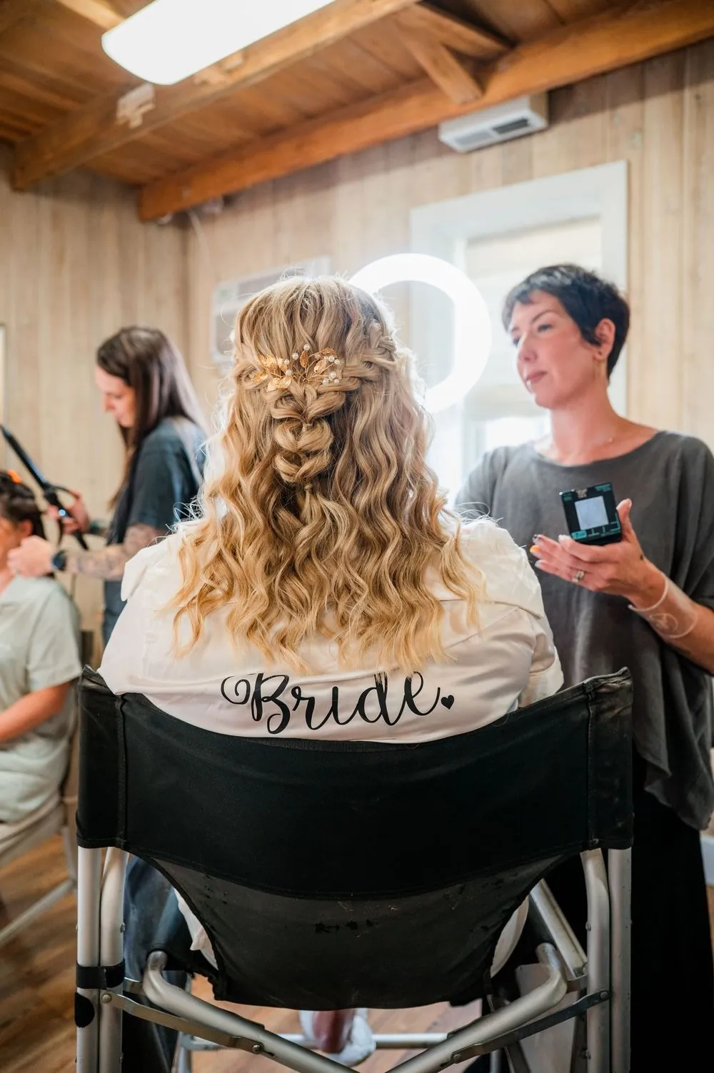 The bride sits in a chair lettered Bride as a stylist curls her blonde hair