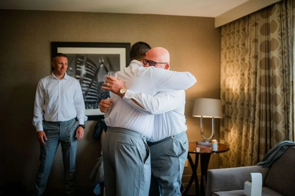 The groom shares a long hug with a bald man in glasses in a hotel room