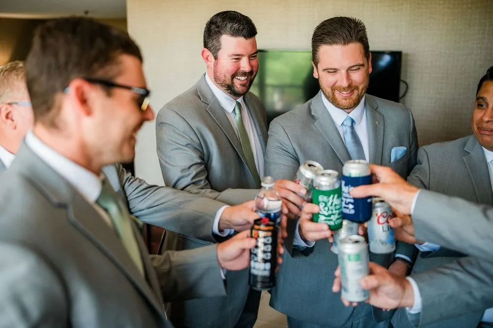 Groomsmen in light gray suits clink beer cans together while getting ready