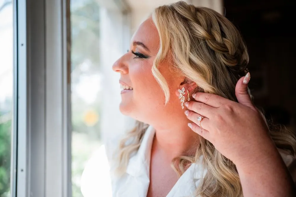 The bride fastens an earring in soft window light, her braid pinned back