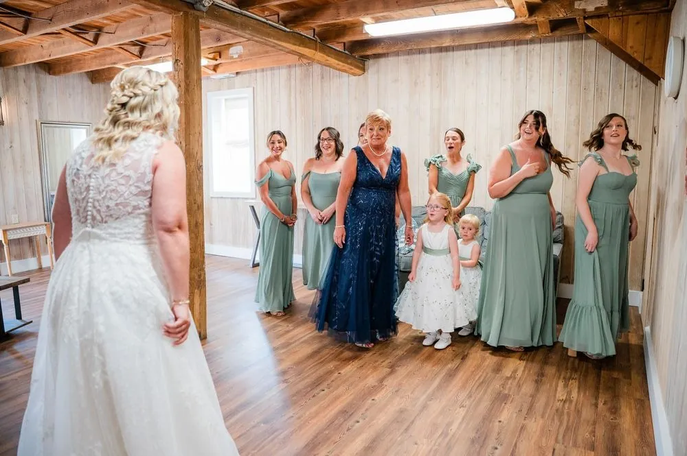 Bridesmaids in sage gowns see the bride in her dress for the first time inside the barn