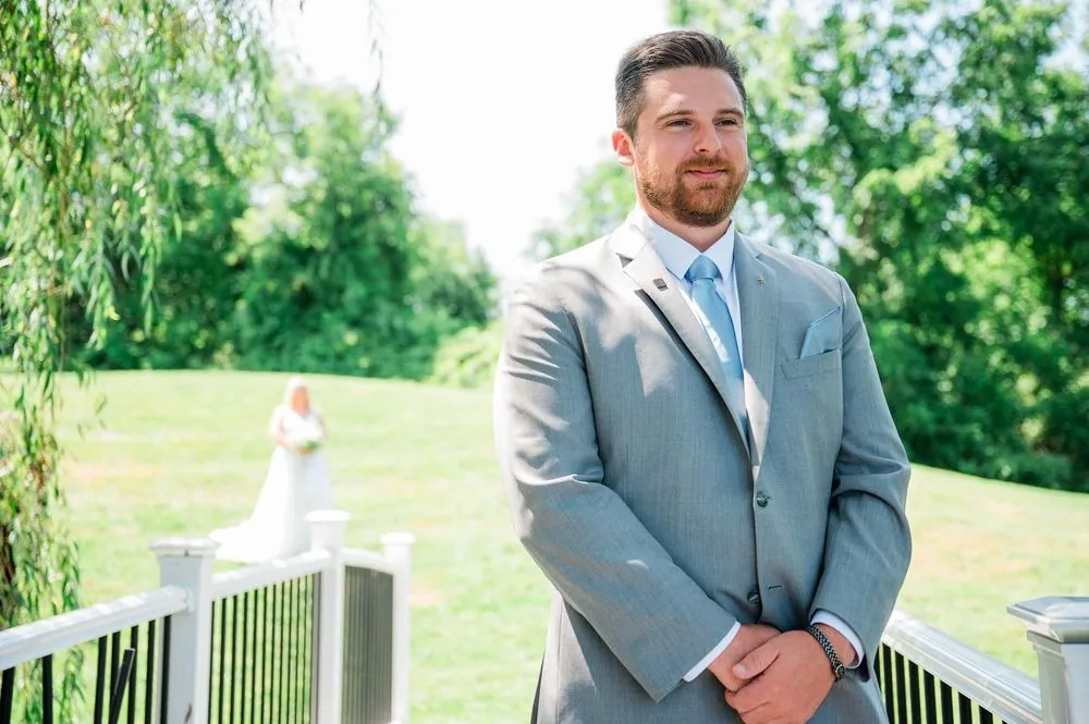 The groom waits on a deck railing, green trees behind him, before seeing the bride