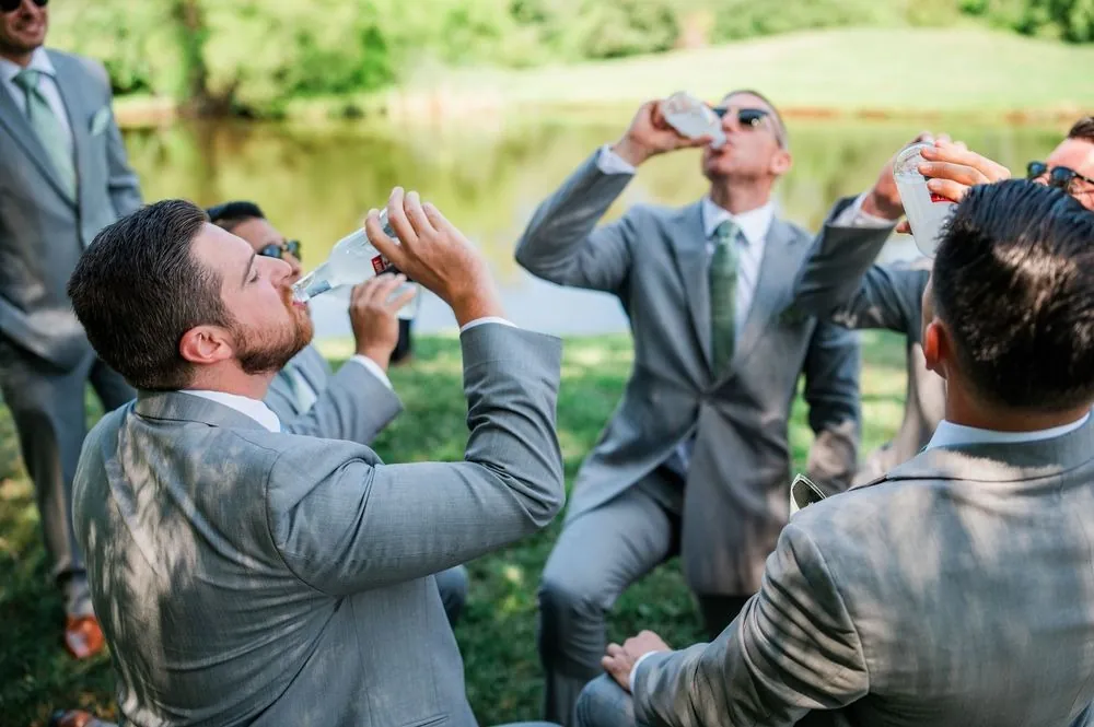 Groomsmen in sunglasses kneel on the lawn and chug bottled drinks by the pond