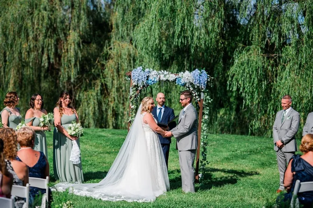 The couple faces each other under a hydrangea arch with the wedding party beneath willows