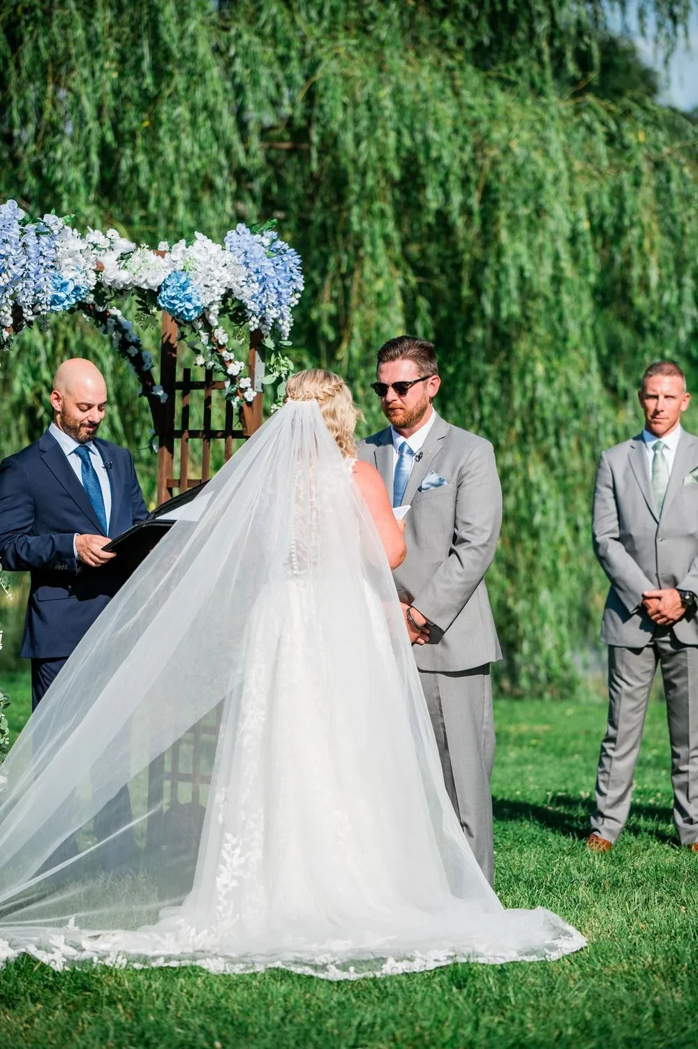 The bride's long cathedral veil spreads across the grass during the vows