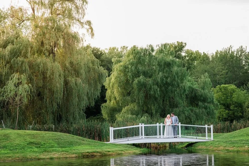 Arianne and Michael stand on a white footbridge over the pond, framed by weeping willows