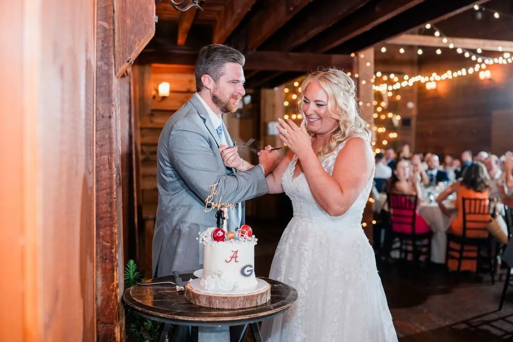 Arianne and Michael laugh while cutting their cake under the barn's string lights