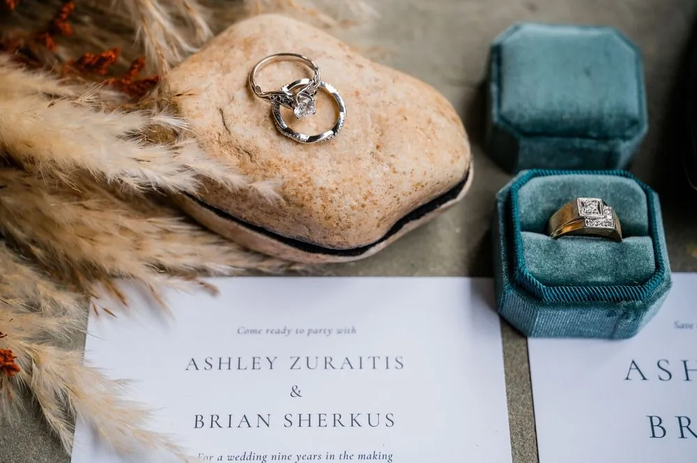 Wedding bands on a stone beside a teal velvet ring box, dried pampas and the printed invitation
