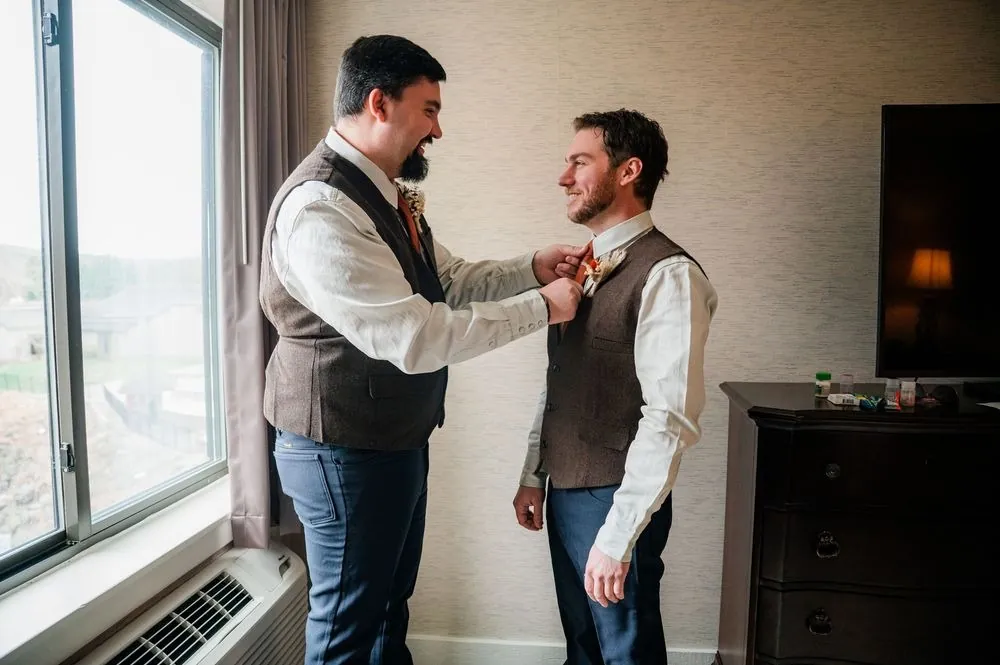 A groomsman in a tweed vest helps the groom with his collar beside the hotel window