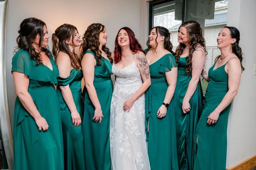 Bridesmaids in forest green gowns gather around the bride in her lace dress in the suite
