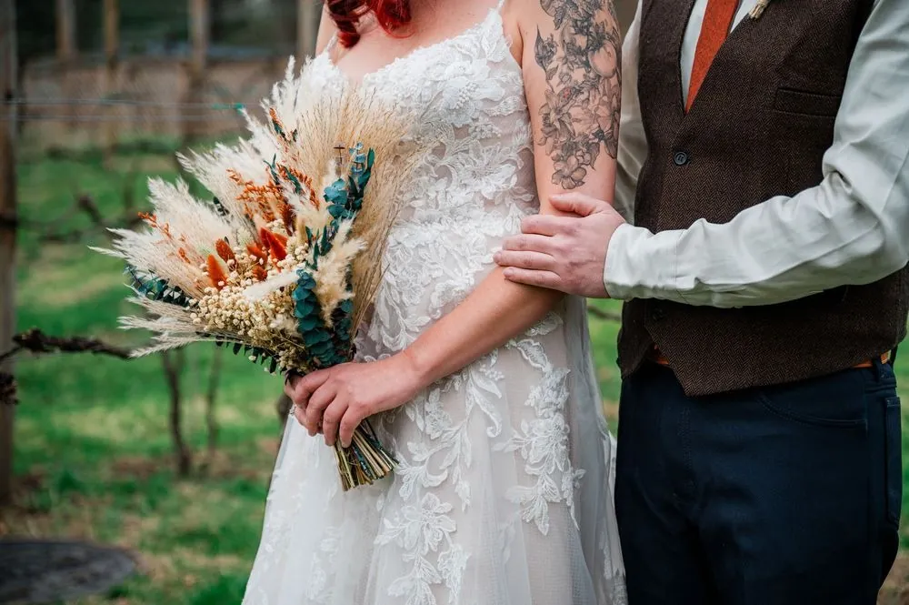 Close crop of the bride's pampas bouquet and tattooed arm linked with the groom's
