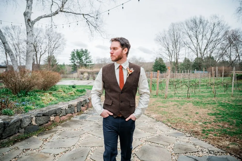 The groom stands on a stone path with hands in pockets, bare trees behind him