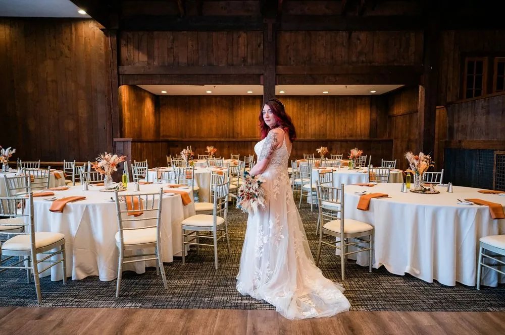 The bride stands alone in the dark timber reception hall among set tables