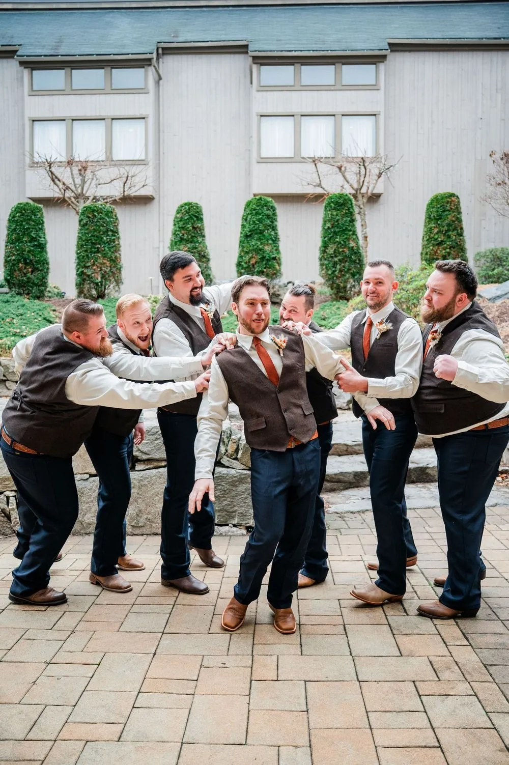 Groomsmen in tweed vests playfully grab the groom on a brick courtyard
