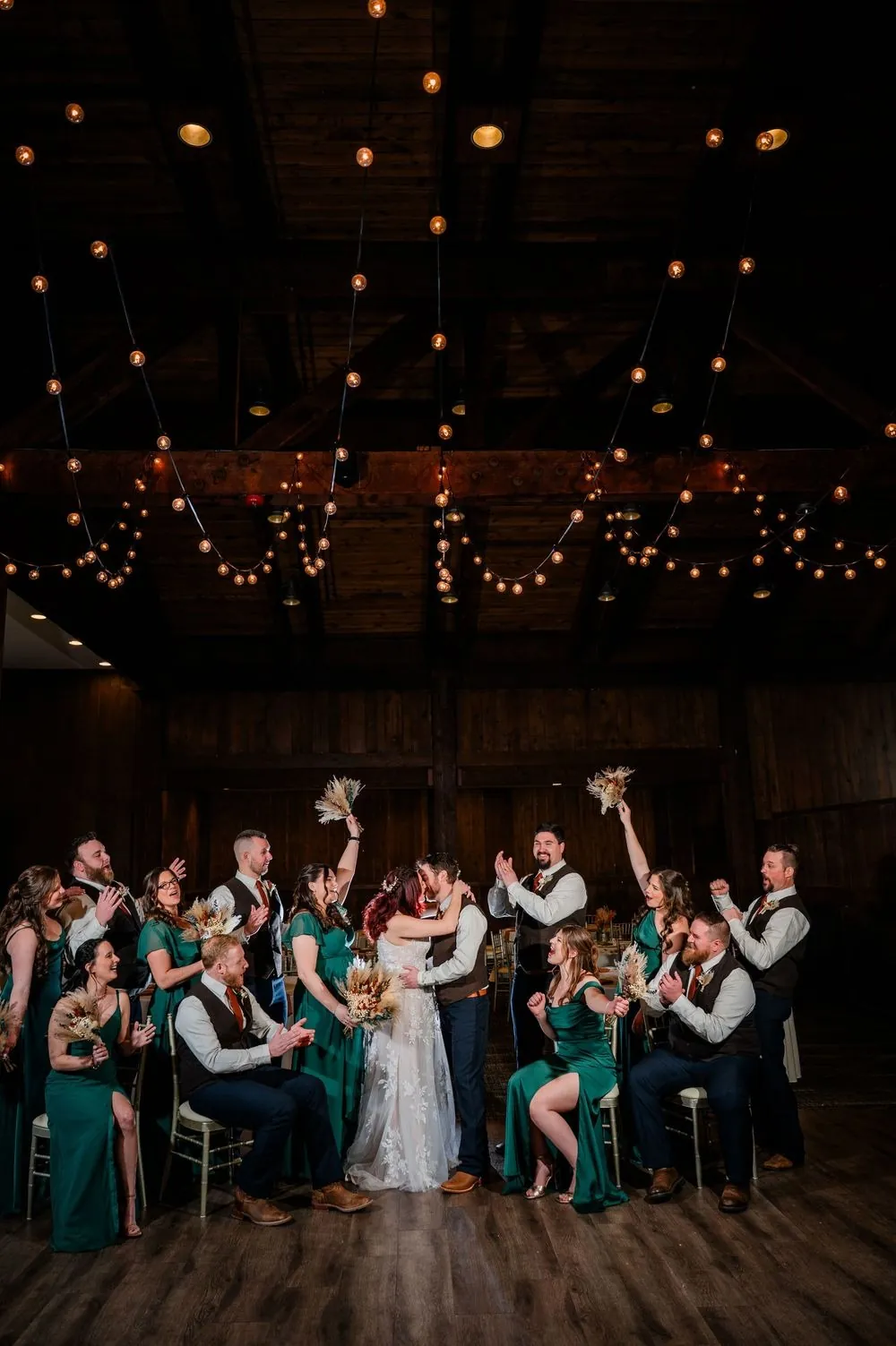 The full wedding party cheers under string lights in the timber hall