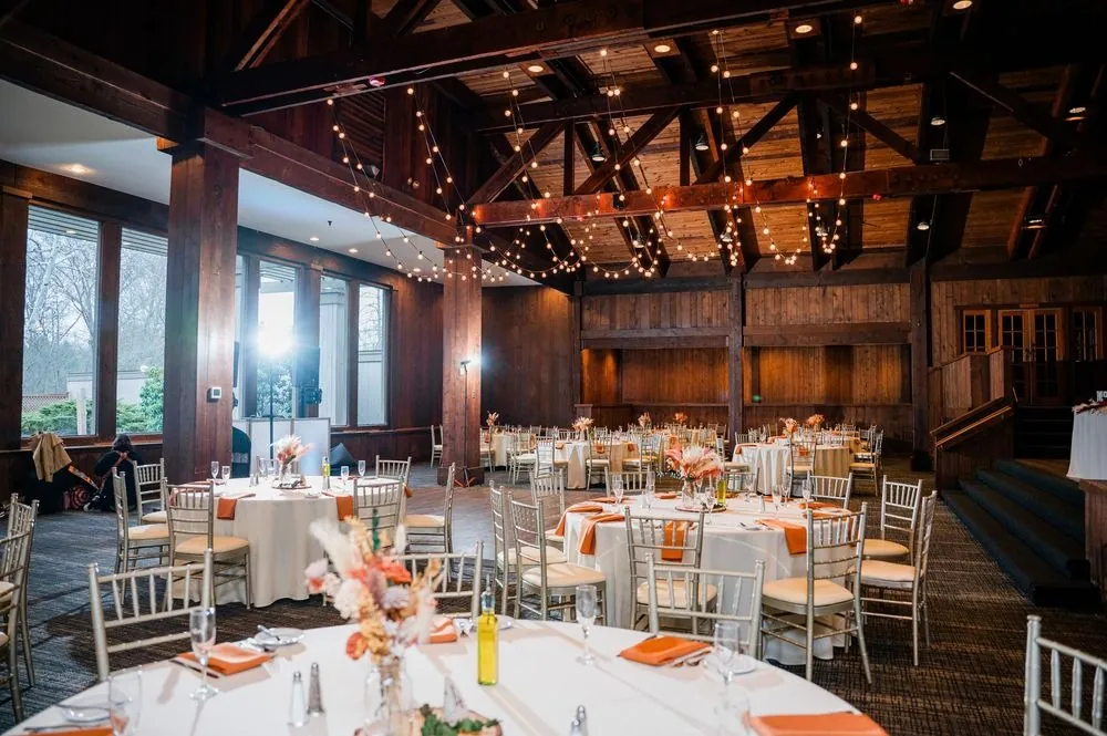 Reception tables with white linens and rust napkins beneath exposed beams and string lights