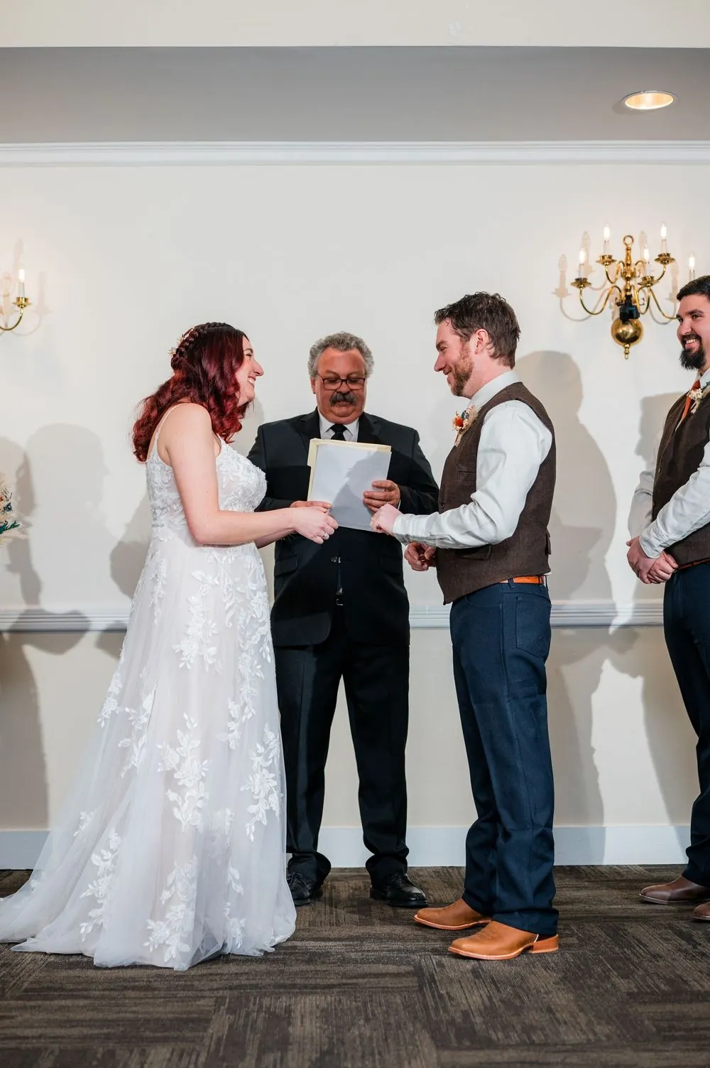 The couple faces the officiant in a cream room with brass candle sconces