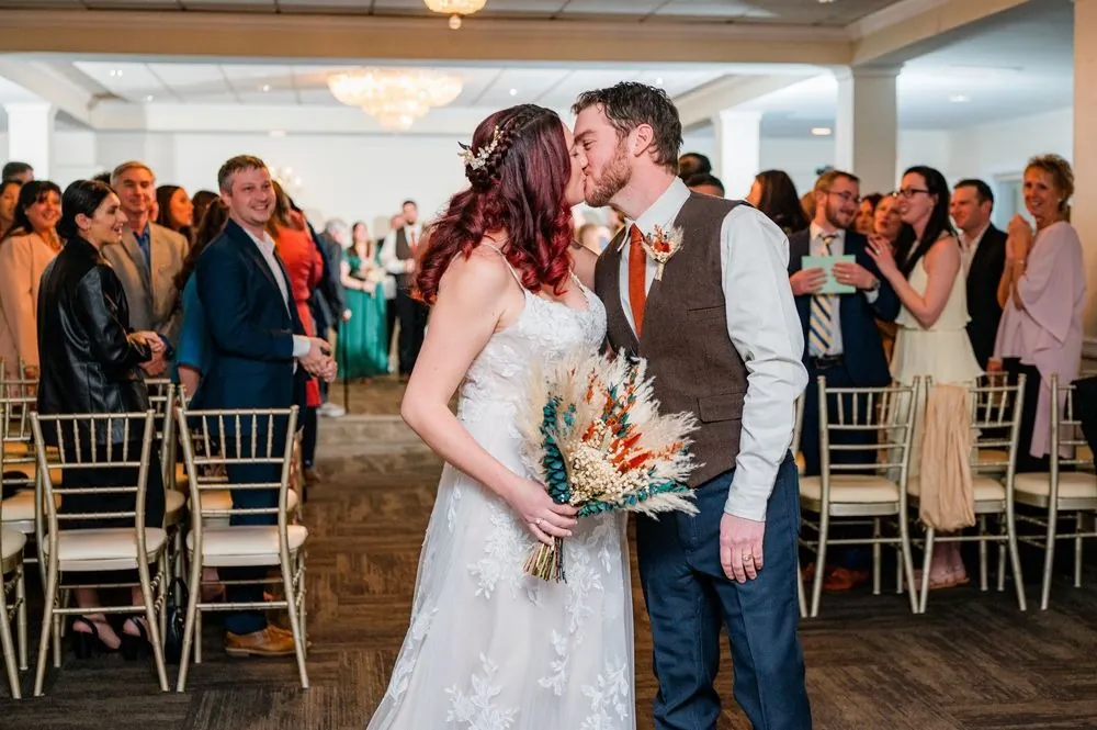 Ashley and Brian kiss in the aisle as seated guests applaud around them