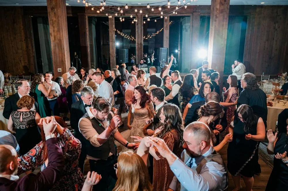 A packed dance floor of guests with arms raised under the timber hall's lights