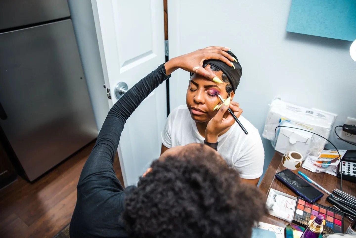 A makeup artist works on the bride's eyebrows at a desk by the window