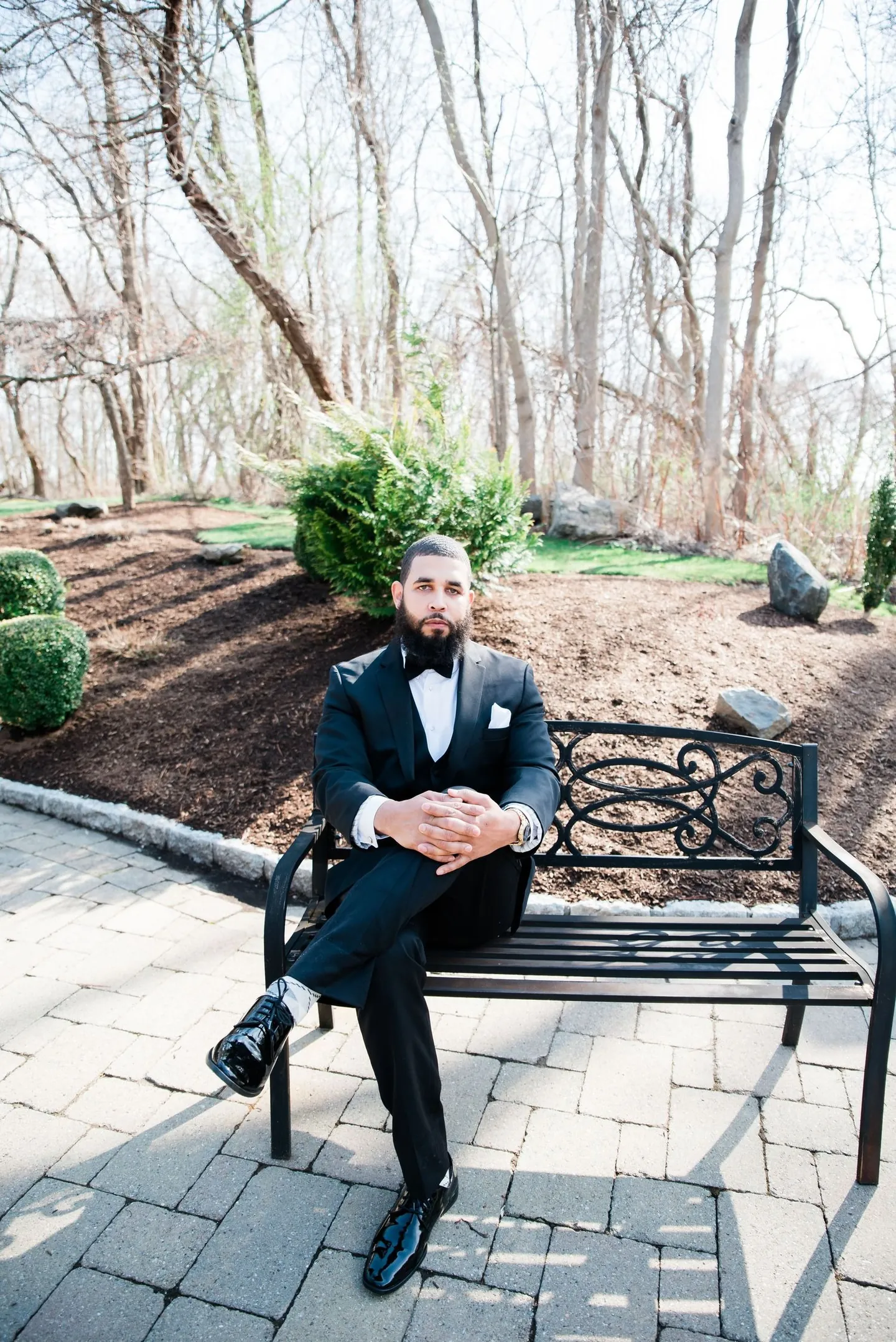 The groom sits on a garden bench in his black tuxedo, legs crossed, before the ceremony