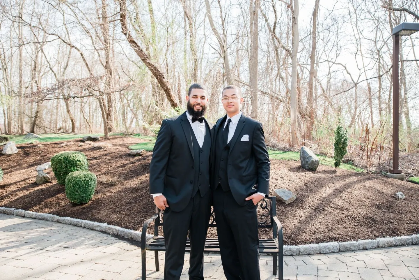 The groom and a groomsman in black tuxedos stand together on a stone garden path