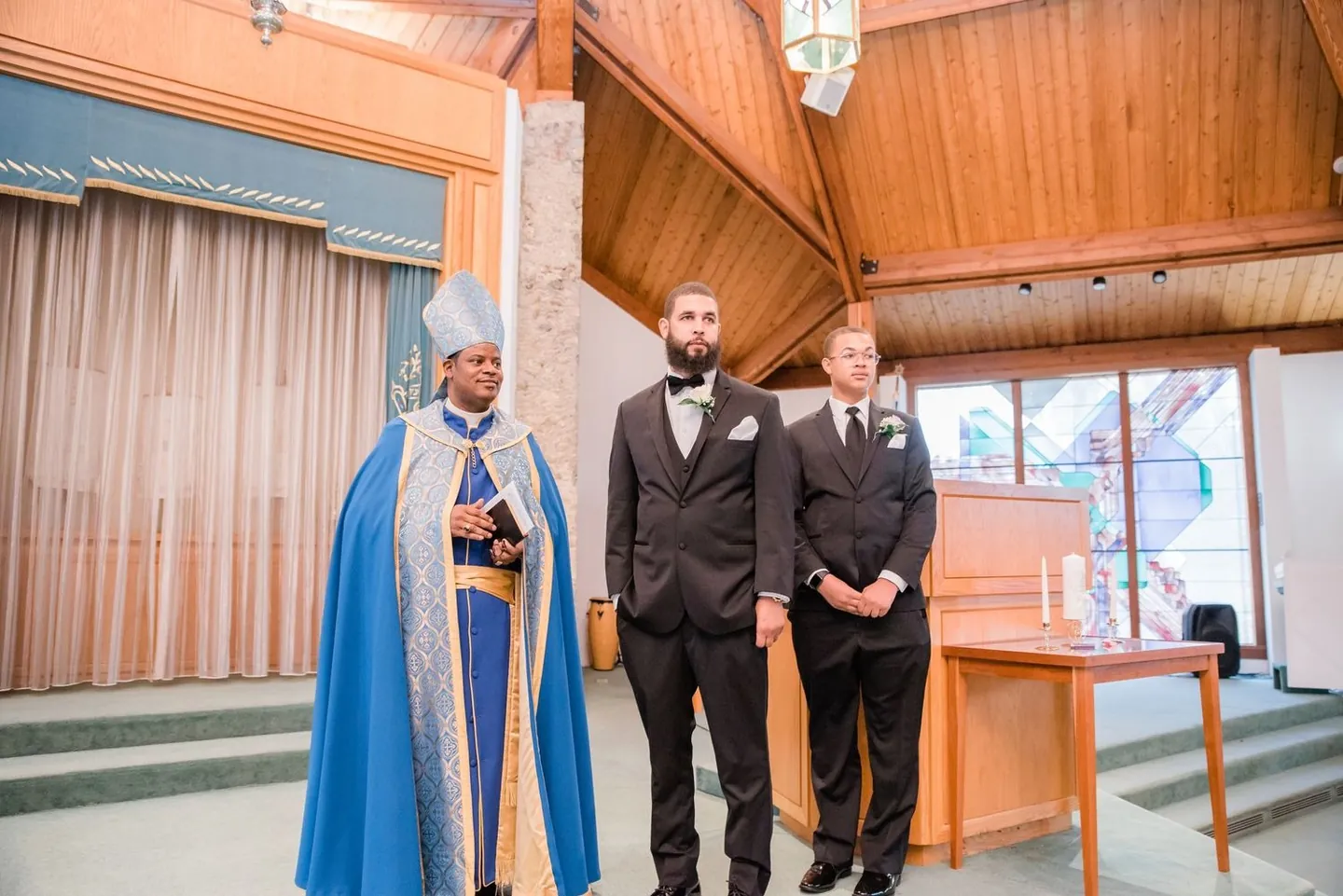 The groom waits at the altar beside an officiant in a mitre and blue and gold vestments