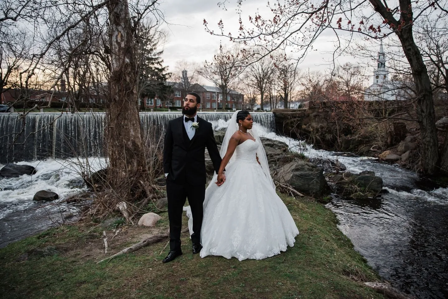 Ashley and Darren hold hands on a grassy point between a waterfall and a rushing stream