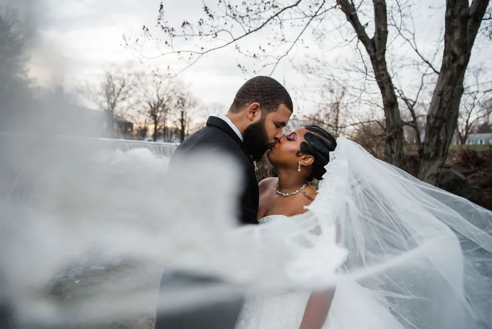 Darren kisses Ashley as her veil blows across the frame by the water