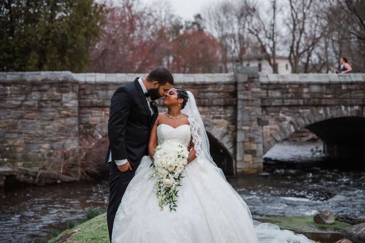 Ashley and Darren kiss in front of an arched stone bridge over the stream