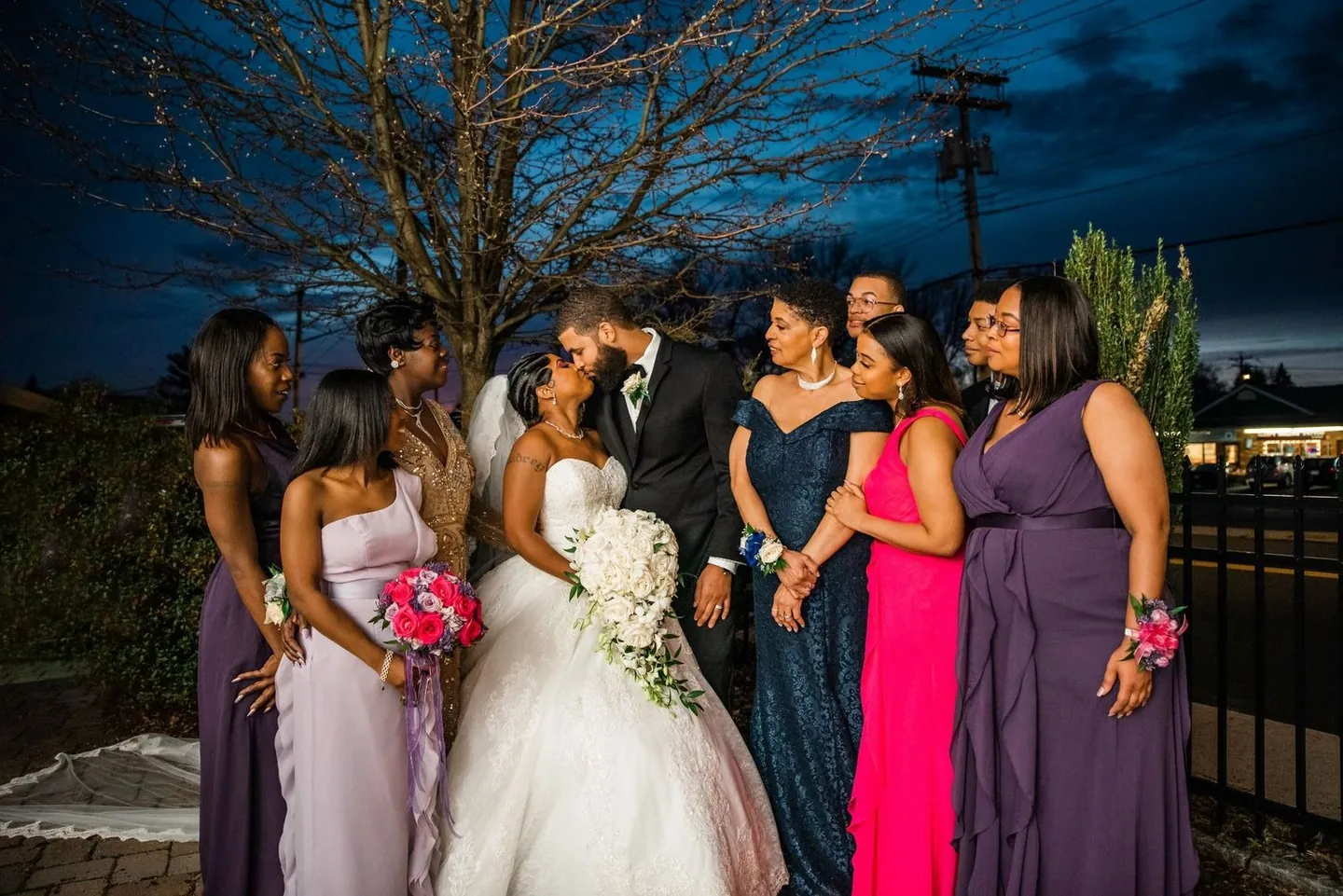 The bride kisses the groom as bridesmaids in purple and pink gowns gather close at dusk